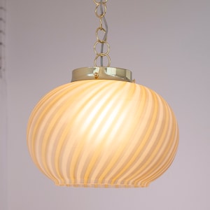 May include: A hanging light fixture with a gold-colored metal canopy and chain. The light has a round, glass shade with a swirl pattern in shades of cream and white. The light is suspended from the ceiling.