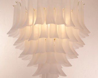 May include: A tiered, white chandelier with a gold-colored chain and ceiling mount. The chandelier features multiple layers of petal-shaped, frosted glass shades, creating a soft, diffused light. The fixture hangs against a white wall.