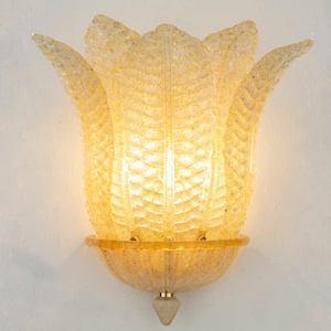 May include: A gold-toned wall sconce with a clear glass shade shaped like a flower. The shade has a textured surface and is illuminated from within.