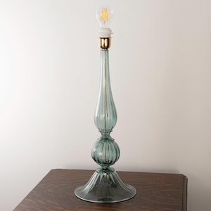 May include: A tall table lamp base made of translucent seafoam green glass with a ribbed, multi-tiered silhouette. The lamp features a gold-toned metal socket at the top, topped with a clear glass LED filament light bulb, creating a slender profile.