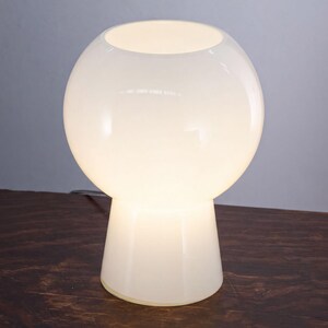A white, round table lamp with a white shade sits on a wooden surface.
