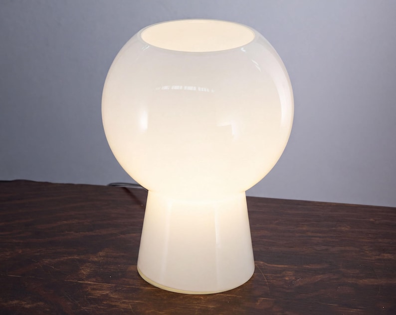 A white, round table lamp with a white shade sits on a wooden surface.