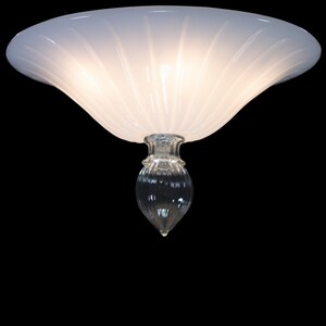 A white, dome-shaped light fixture with a clear glass shade and a pointed finial at the bottom, illuminated and casting a warm glow against a dark background.