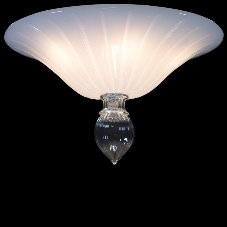 A white, dome-shaped light fixture with a clear glass shade and a pointed finial at the bottom, illuminated and casting a warm glow against a dark background.