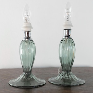 Op de afbeelding: Een paar tafellampen met saliegroene, verticaal geribbelde glazen voetstukken met een brede, uitlopende ronde voet. Elke lamp heeft een hals van gepolijst chroom, een witte kunststof fitting en een heldere, vlamvormige gloeilamp.