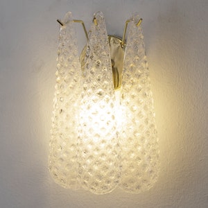 Può includere: Un'applique da parete in vetro trasparente con una superficie testurizzata e una cornice in metallo dorato. L'applique ha tre lampadine accese, che proiettano una luce calda.