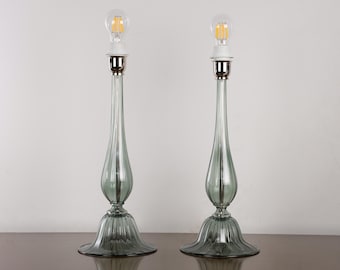 Può includere: Una coppia di lampade da tavolo alte con basi in vetro scanalato grigio fumo traslucido, caratterizzate da una base svasata a forma di campana e un collo sottile e allungato. Ogni lampada ha un portalampada in metallo argentato e una lampadina LED.