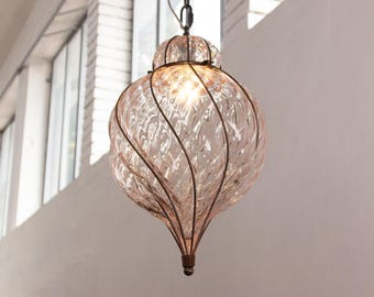 May include: A teardrop-shaped pendant light fixture featuring a textured, pale pink glass shade encased in a dark metal wire cage with a spiral design. The light is illuminated from within, casting a warm glow through the rippled glass surface.