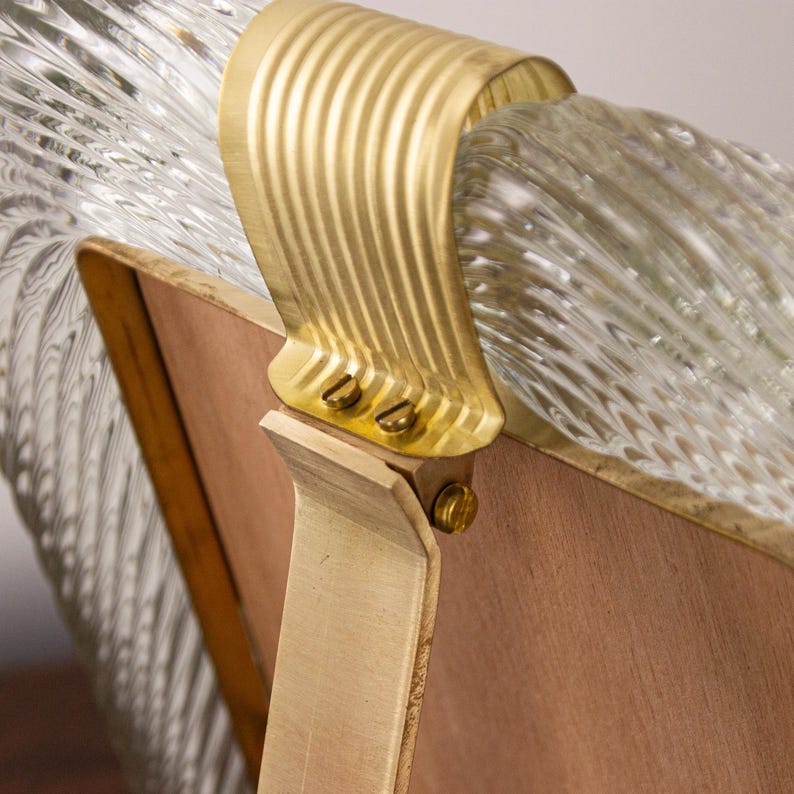 May include: A vintage picture frame with a clear glass front and a gold metal frame. The frame has a unique design with a wavy, textured glass front. The back of the frame is made of wood and has a gold metal stand.