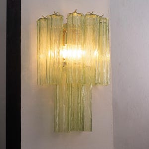 Peut inclure: Un luminaire mural avec un design en couches. Le luminaire présente des panneaux de verre vert pâle verticaux et nervurés, avec une lumière chaude émanant de l'intérieur. Le luminaire est monté sur un mur blanc.