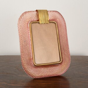 Op de afbeelding: Een roze glazen fotolijst met een gouden metalen rand. De lijst heeft een getextureerd oppervlak en een rechthoekige opening voor een foto.