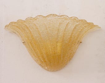 May include: A semi-circular wall sconce made of textured, amber-colored glass. The fixture features a scalloped, fan-like shape with vertical ridges radiating from the base. Small gold-toned metal hardware secures the glass to the wall mount.