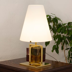 May include: A gold table lamp with a white lampshade and a base made of stacked glass cubes. The lamp is on a dark wood table.