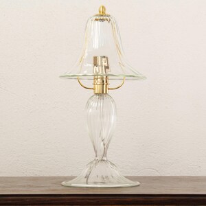 A clear glass lamp with a gold-colored base and a clear glass shade is placed on a wooden surface against a white wall.