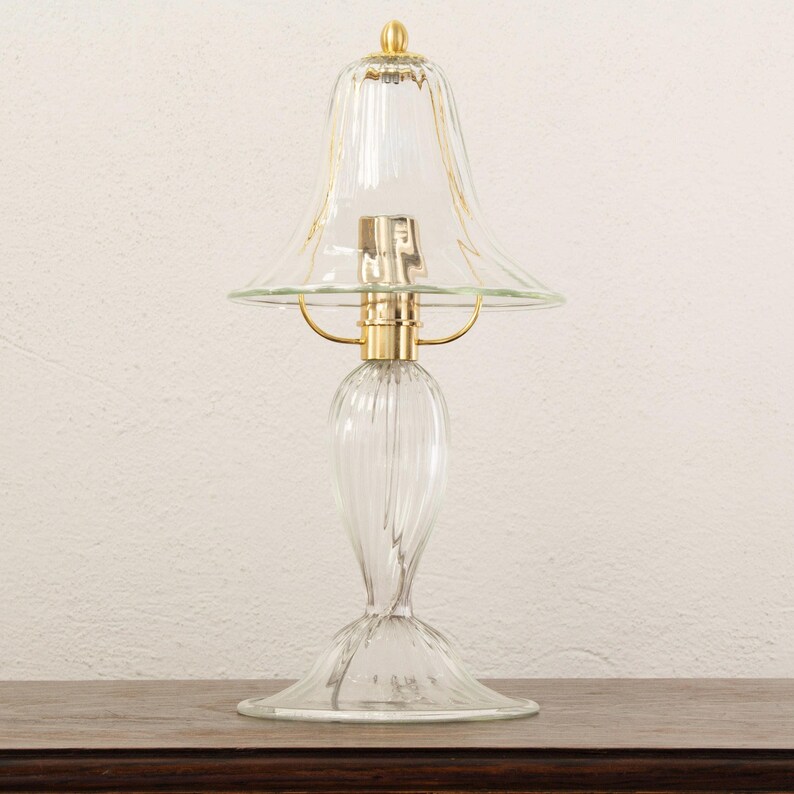 A clear glass lamp with a gold-colored base and a clear glass shade is placed on a wooden surface against a white wall.