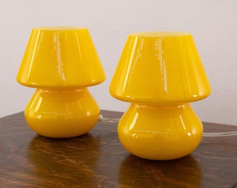 May include: A pair of matching table lamps with a glossy yellow finish. Each lamp features a rounded base and a wide, flared shade in a singular mushroom-like silhouette. The lamps are constructed from a smooth, reflective material with clear cords.