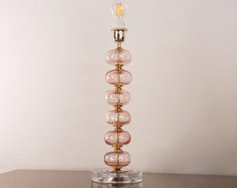 May include: A tall table lamp featuring a central gold-toned metal rod stacked with six translucent, pale pink, donut-shaped glass spheres. The lamp stands on a clear, circular glass base and is topped with a single, exposed filament light bulb.