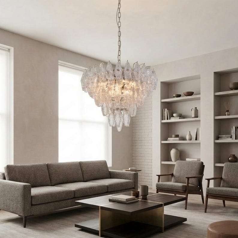 a modern living room with a large crystal chandelier hanging from the ceiling, casting a warm glow throughout the space. The room is furnished with a gray sofa, two armchairs, and a coffee table. The walls are painted in a light color, and the room is well-lit by natural light coming through a window.