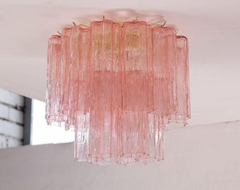 Può includere: Un lampadario a più livelli in vetro rosa con una montatura in metallo color oro. Il vetro ha un design verticale strutturato. L'apparecchio è montato su un soffitto bianco.