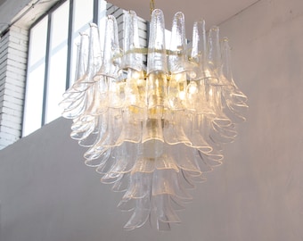 May include: A clear glass chandelier with a tiered, floral-like design. The chandelier has multiple layers of petal-shaped glass elements, creating a cascading effect. The fixture is suspended from a gold-colored metal frame and chain, and is lit from within.