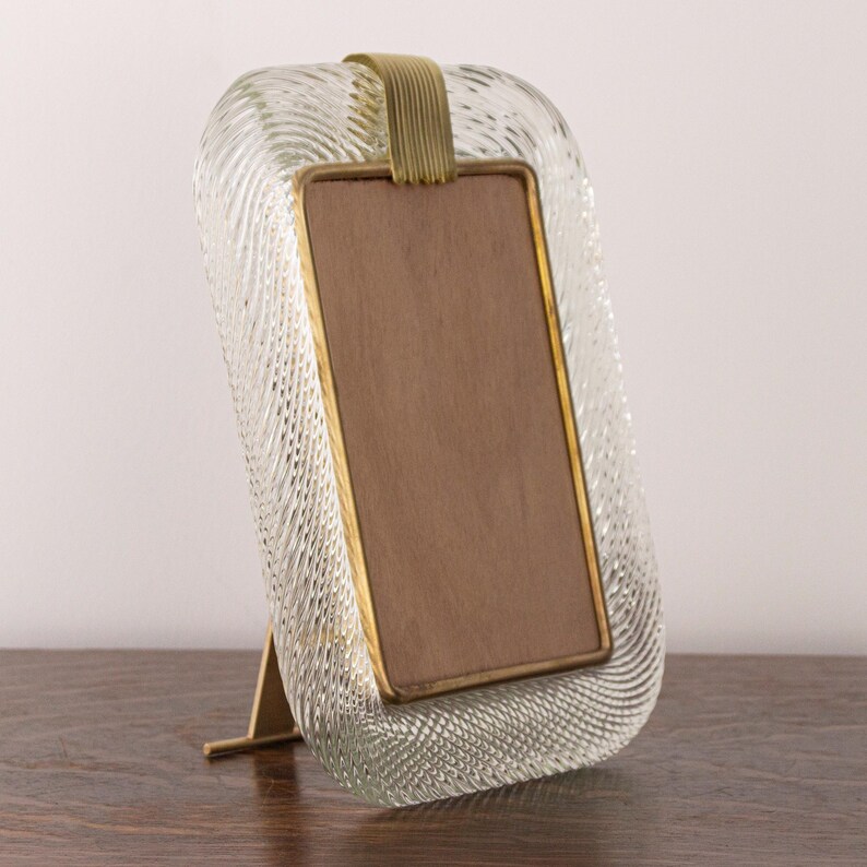 May include: A clear glass picture frame with a textured surface and a brass stand. The frame has a brass trim and a brown backing.