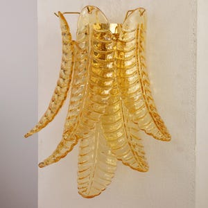 May include: A wall sconce featuring multiple amber-coloured glass panels shaped like elongated leaves. The translucent glass pieces are arranged in a layered, fan-like formation, attached to a central gold-toned metal mounting bracket.