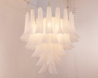 May include: A white, tiered chandelier with a floral design. The light fixture is suspended from a gold chain and features multiple layers of frosted, petal-shaped shades. The chandelier is illuminated, casting a soft glow.