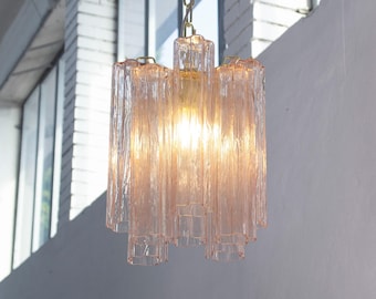 Puede incluir: Una lámpara colgante con múltiples varillas de vidrio verticales, texturizadas y de color rosa pálido, dispuestas en un grupo cilíndrico. La luz está suspendida por una cadena de metal dorado y emite un brillo cálido desde el centro del vidrio.