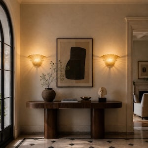 a well-lit hallway with a wooden console table, a framed abstract artwork, and two wall sconces. The console table has a vase with a plant and a bowl on it. The hallway also has a fireplace and a doorway leading to another room.