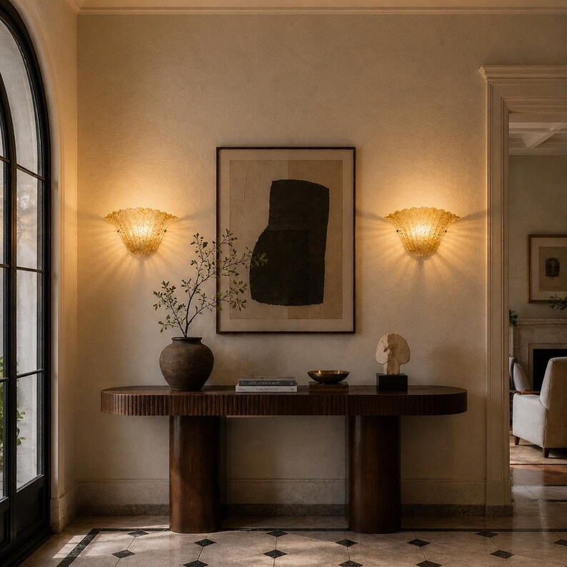 a well-lit hallway with a wooden console table, a framed abstract artwork, and two wall sconces. The console table has a vase with a plant and a bowl on it. The hallway also has a fireplace and a doorway leading to another room.