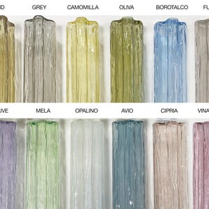 a collection of twelve glass vases, each with a unique color and pattern, arranged in two rows of six. The vases are labeled with their names, such as Indigo, Sand, Grey, Camomilla, Olive, Borotalco, Fume, True Pink, Ralto, Malve, Mela, Opalino, Avio, Cipri, Vinacia, and Champagne.