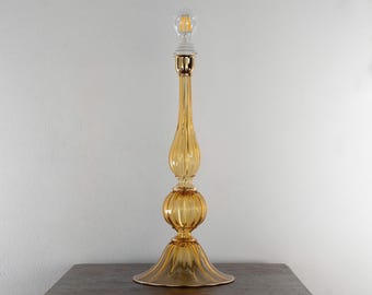 May include: A tall table lamp featuring a translucent amber-colored glass base with a ribbed, multi-tiered silhouette. The lamp includes a gold-toned metal neck and a clear glass light bulb at the top, creating a slender and elongated vertical form.