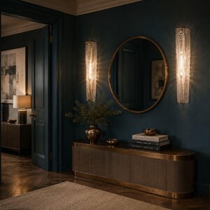 a luxurious and elegant hallway with dark blue walls, a large oval mirror, and two crystal chandeliers hanging above the mirror. The hallway features a wooden console table with a vase of greenery and a stack of books, creating a sophisticated and inviting atmosphere.