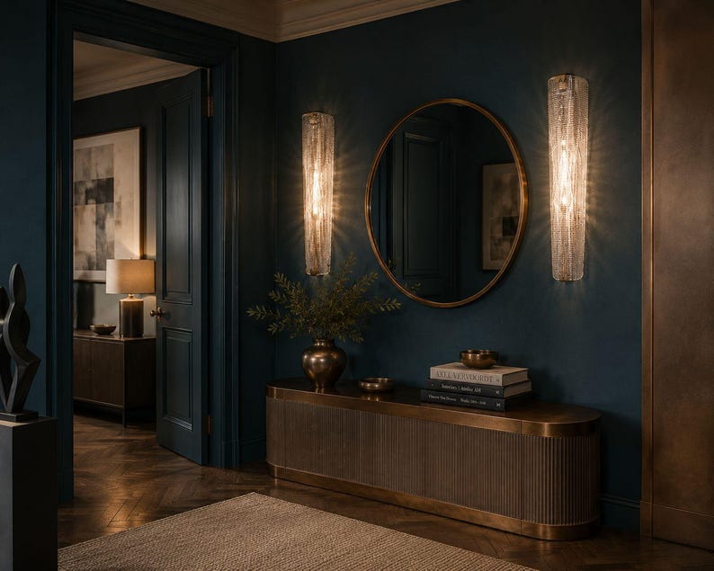 a luxurious and elegant hallway with dark blue walls, a large oval mirror, and two crystal chandeliers hanging above the mirror. The hallway features a wooden console table with a vase of greenery and a stack of books, creating a sophisticated and inviting atmosphere.