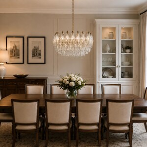a formal dining room with a large wooden table surrounded by six matching chairs. A crystal chandelier hangs above the table, casting a warm glow over the space. The walls are painted in a light color, and there are framed pictures on the walls. A vase of flowers sits on the table, adding a touch of natural beauty to the elegant setting.