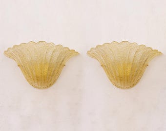 May include: A pair of wall sconces featuring a fan-shaped design made of textured, translucent yellow glass. Each sconce has a scalloped edge and a ribbed surface pattern, with small metal hardware visible on the sides for mounting purposes.