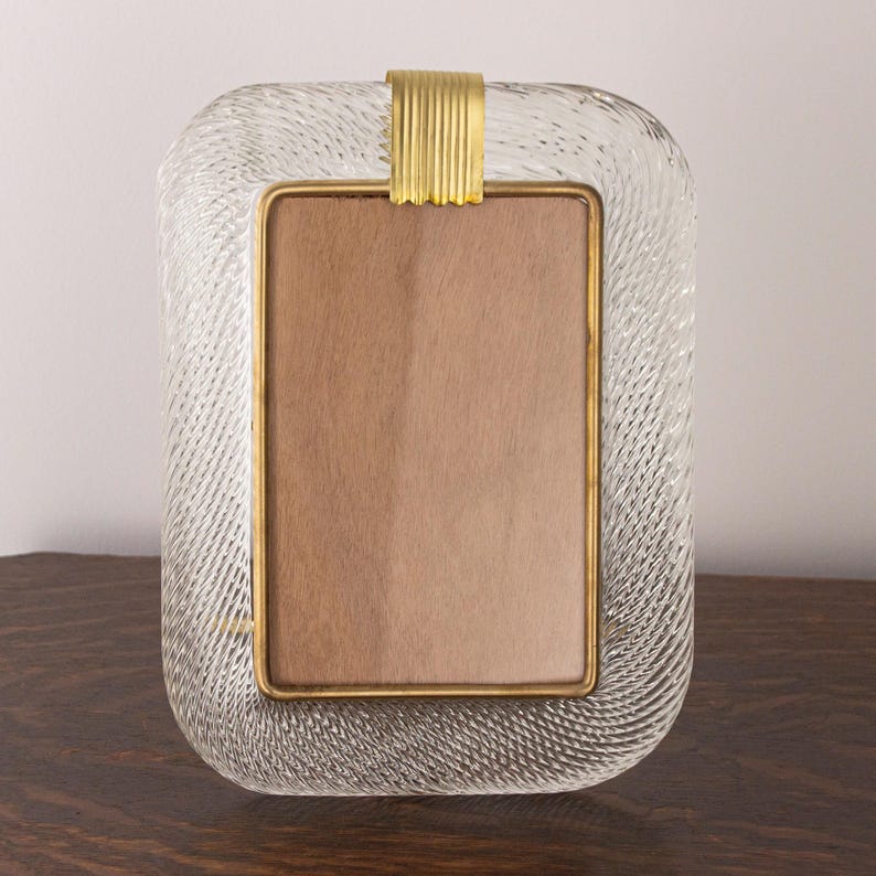 May include: A clear glass picture frame with a gold metal trim and a textured surface. The frame has a rectangular shape with rounded corners.