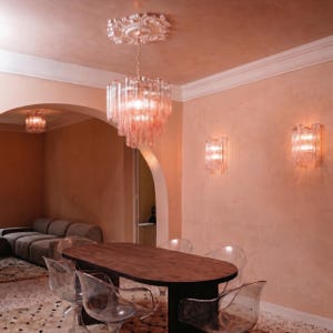 a modern dining room with a wooden table and clear glass chairs. The room has a pink and peach color scheme, with a chandelier hanging from the ceiling and wall sconces providing lighting.