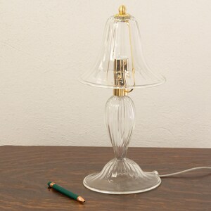 A clear glass lamp with a gold-colored base and a green pen on a wooden surface.