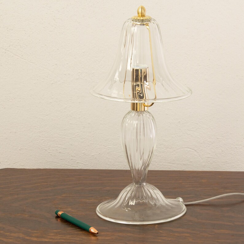 A clear glass lamp with a gold-colored base and a green pen on a wooden surface.