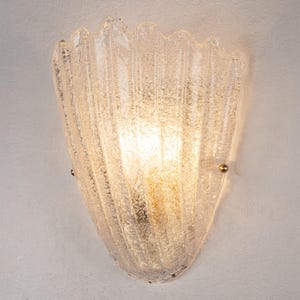 Op de afbeelding: Een wandlamp met een halfronde, getextureerde glazen kap met verticale ribbels. Het glas heeft een ruwe, matachtige uitstraling en is aan de muur bevestigd met kleine, ronde metalen bevestigingspunten aan de linker- en rechterkant.