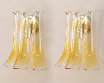 May include: A pair of wall sconces featuring three vertical, curved glass panels with a clear and amber gradient finish. Each fixture is mounted on a circular, textured gold-tone metal base that supports the glass elements in a fan-like arrangement.