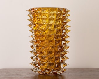 May include: A cylindrical amber-colored glass vase featuring a prominent spiked, geometric texture across its entire exterior surface. The glass is translucent, showcasing a deep golden hue with sharp, protruding points arranged in a grid pattern.