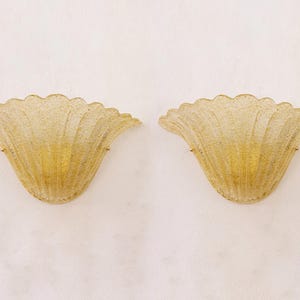 May include: A pair of wall sconces, each with a fan-shaped design. They are made of textured, golden-yellow glass and have gold-coloured metal fixings. The sconces are reminiscent of a seashell.