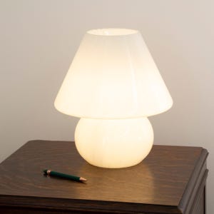May include: A mushroom-shaped table lamp made of white glass with a smooth, glowing finish. The lamp features a wide, conical shade resting on a rounded, bulbous base. The light emits a soft, warm illumination throughout the entire glass structure.