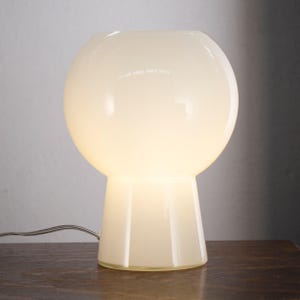 A white, round table lamp with a white shade sits on a wooden surface.