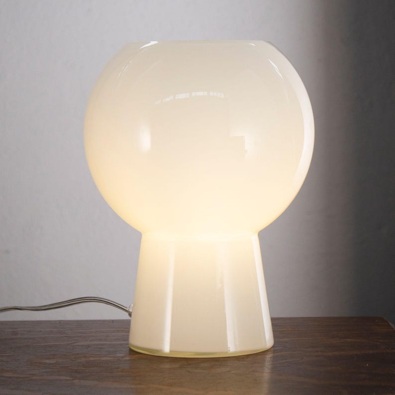 A white, round table lamp with a white shade sits on a wooden surface.