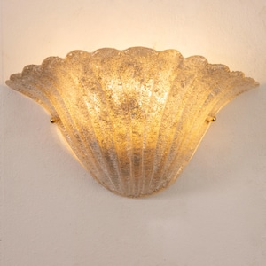May include: A wall-mounted light fixture with a shell-like design. The fixture is made of textured, translucent glass with a golden hue. The light shines through the glass, creating a warm glow. Two small gold-colored accents are visible.