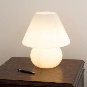 Może przedstawiać: Biała lampa stołowa w kształcie grzyba z białym abażurem. Lampa jest włączona i oświetla pomieszczenie. Lampa stoi na ciemnobrązowym drewnianym stole.