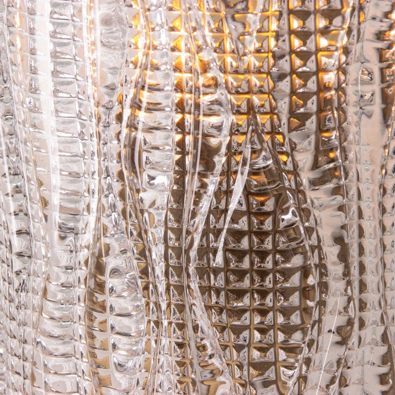 a close-up view of a textured, glass surface with a repeating pattern of small, square-shaped lines and curves. The glass appears to be made of a clear material, possibly glass or a similar transparent material, with a metallic or textured finish. The image does not contain any discernible objects or actions, and the focus is on the intricate and visually interesting pattern created by the glass surface.